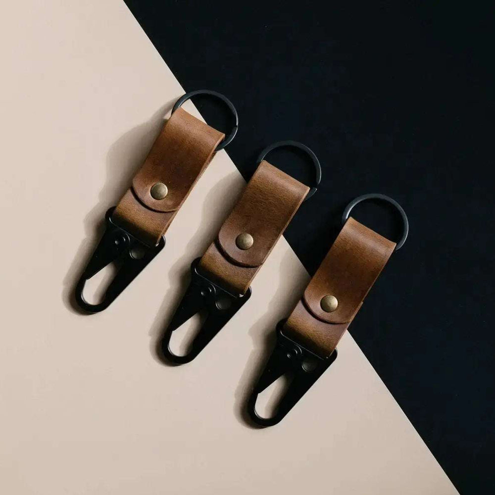 Set of three brown leather keychains with tactical black carabiner clips on contrasting beige and black background