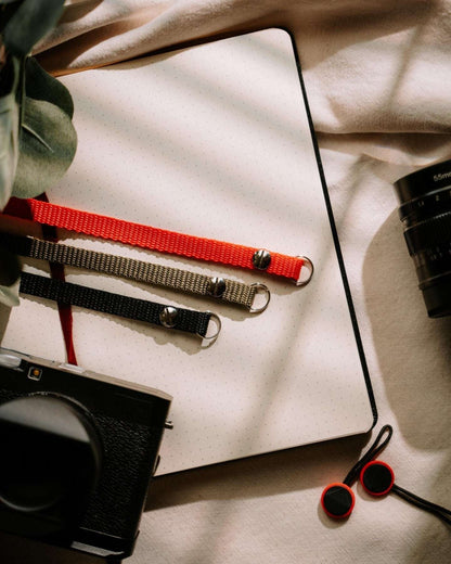 Thin vintage camera straps in red, gray, and black laid on an open notebook with camera accessories nearby