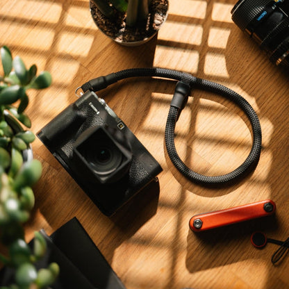 Short black vintage camera strap (Kurzes Basic Kameraseil) attached to a compact camera on a wooden surface with plants and camera accessories nearby