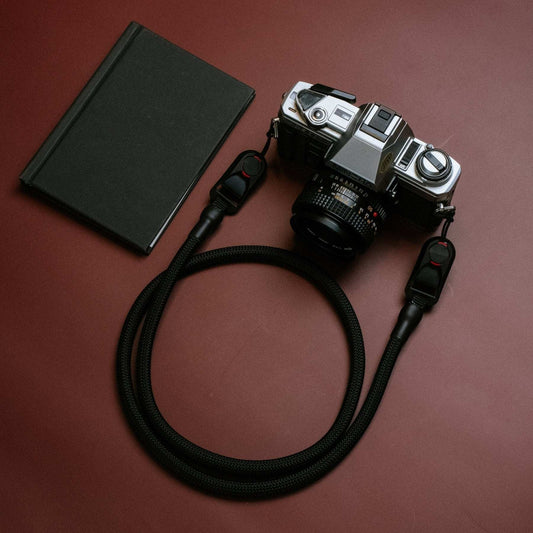Quick Release Kameragurt vintage camera rope strap with durable black polypropylene climbing rope and quick-release adapters attached to an analog camera on a brown background next to a black notebook